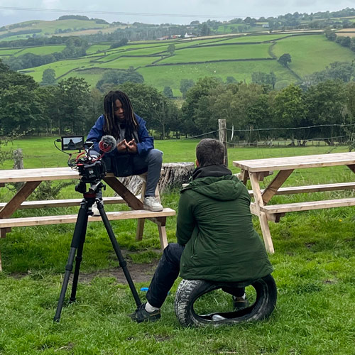 boy chatting to the camera as he's being interviewed about his residential experience. He's sat on a bunch with welsh fields and sheep as his backdrop