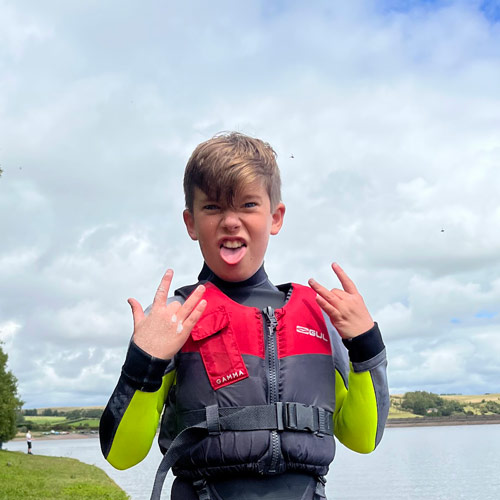 12 year old sticking his tongue out happily at the camera.He's wearing a wetsuit and buoyancy aid as he's about to stand up paddle boarding
